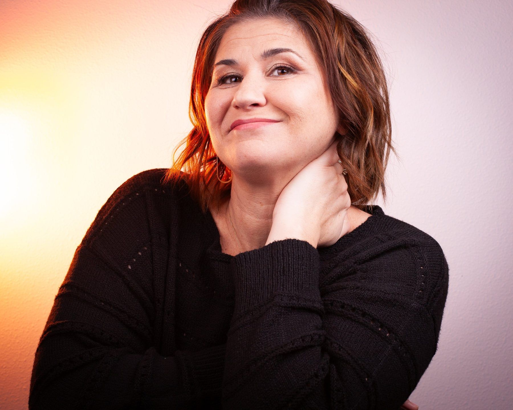 Lori Lackland in a black sweater, smiling, one hand at her neck, with warm amber light falling from the left.