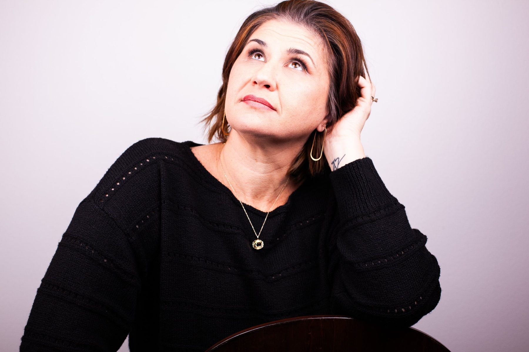 Lori Lackland in a black sweater, one hand in her hair, looking upward in thought.